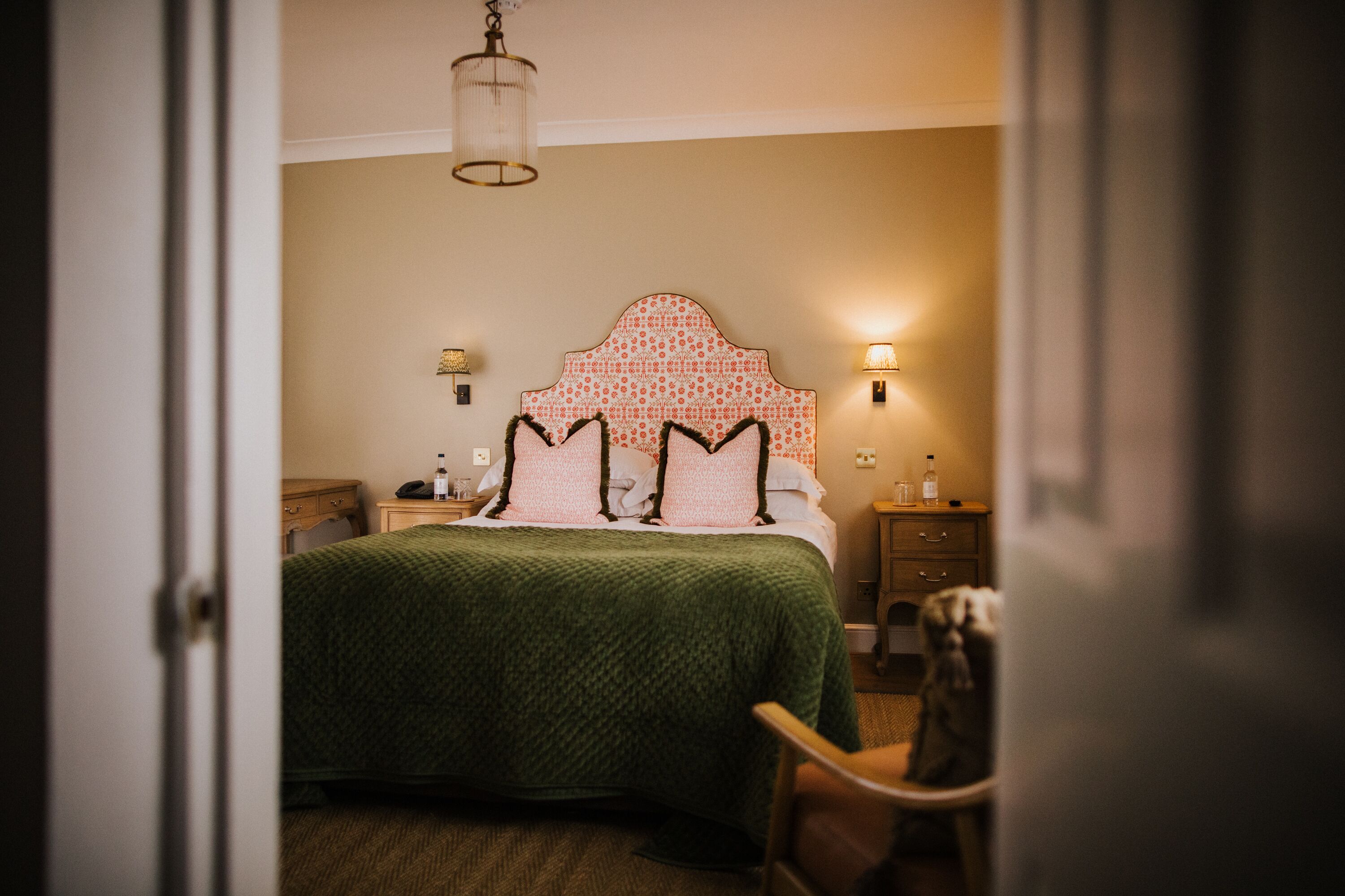 A bedroom with a green bedspread, a patterned headboard, bedside lamps, and a nightstand with a bottle.