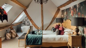 Un dormitorio con techo inclinado, una cama grande con varias almohadas, una mesita de noche con lámpara, una mesita auxiliar de madera y un cuadro en la pared.