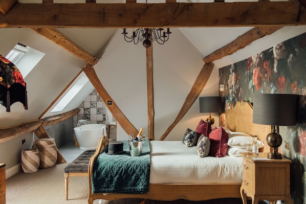 A bedroom with a sloped ceiling, a large bed with multiple pillows, a bedside table with a lamp, a wooden nightstand, and a painting on the wall. - The Farmhouse at Redcoats Restaurant & Hotel (Hitchin)
