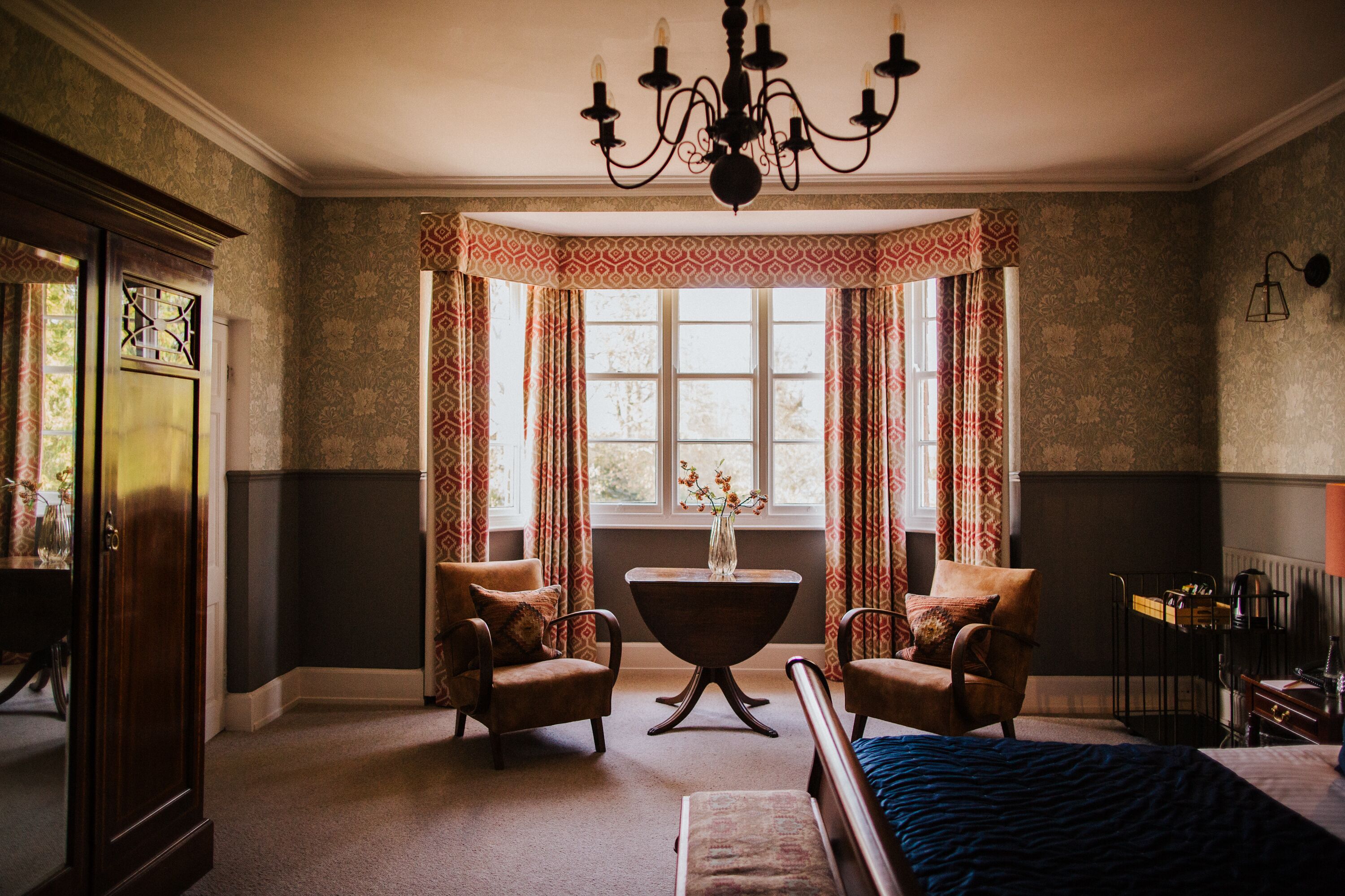 A room with a bed, two armchairs, a table, a chandelier, a window with curtains, and a radiator.