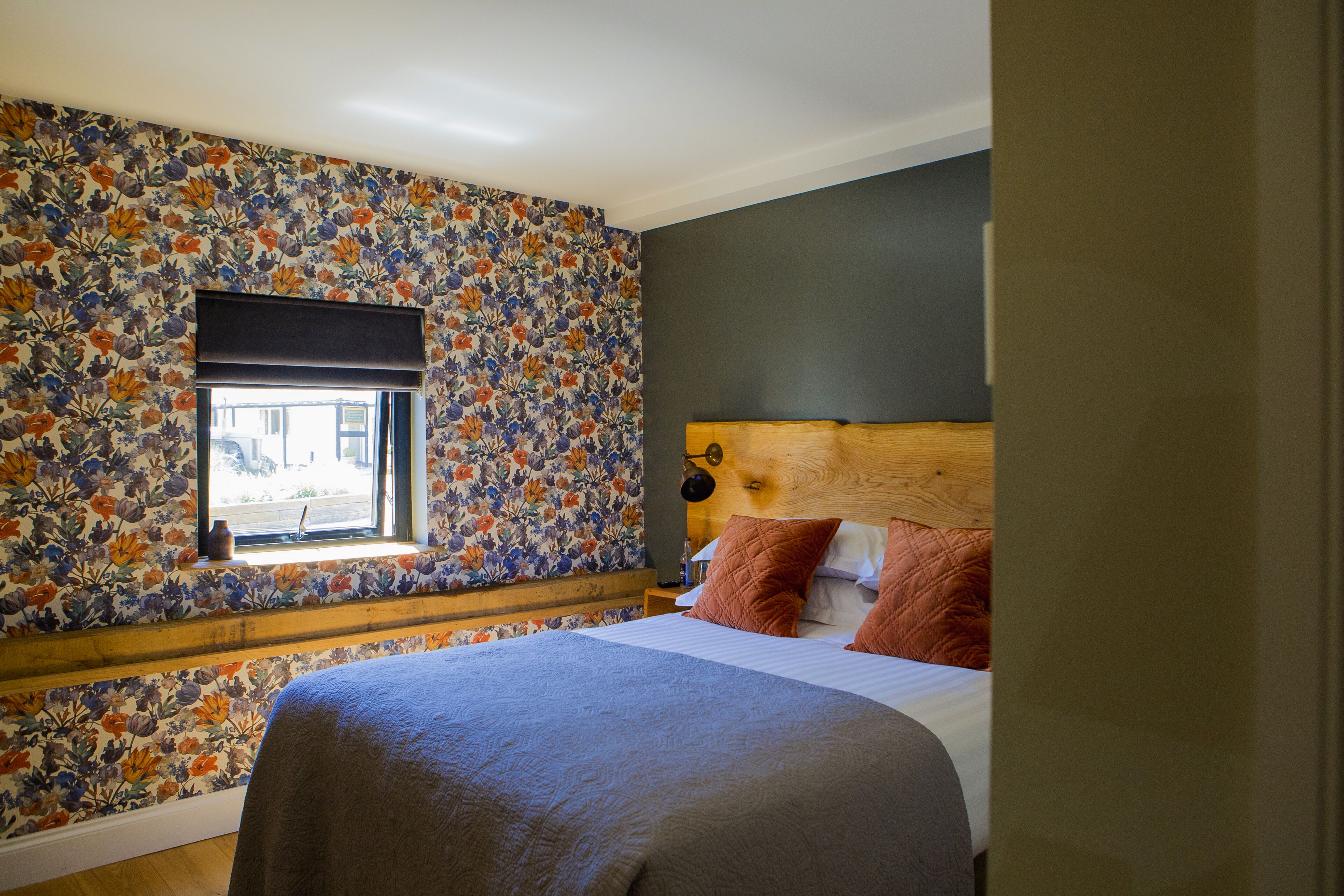 A bedroom with a floral wallpaper, a wooden headboard, and a window with a view.