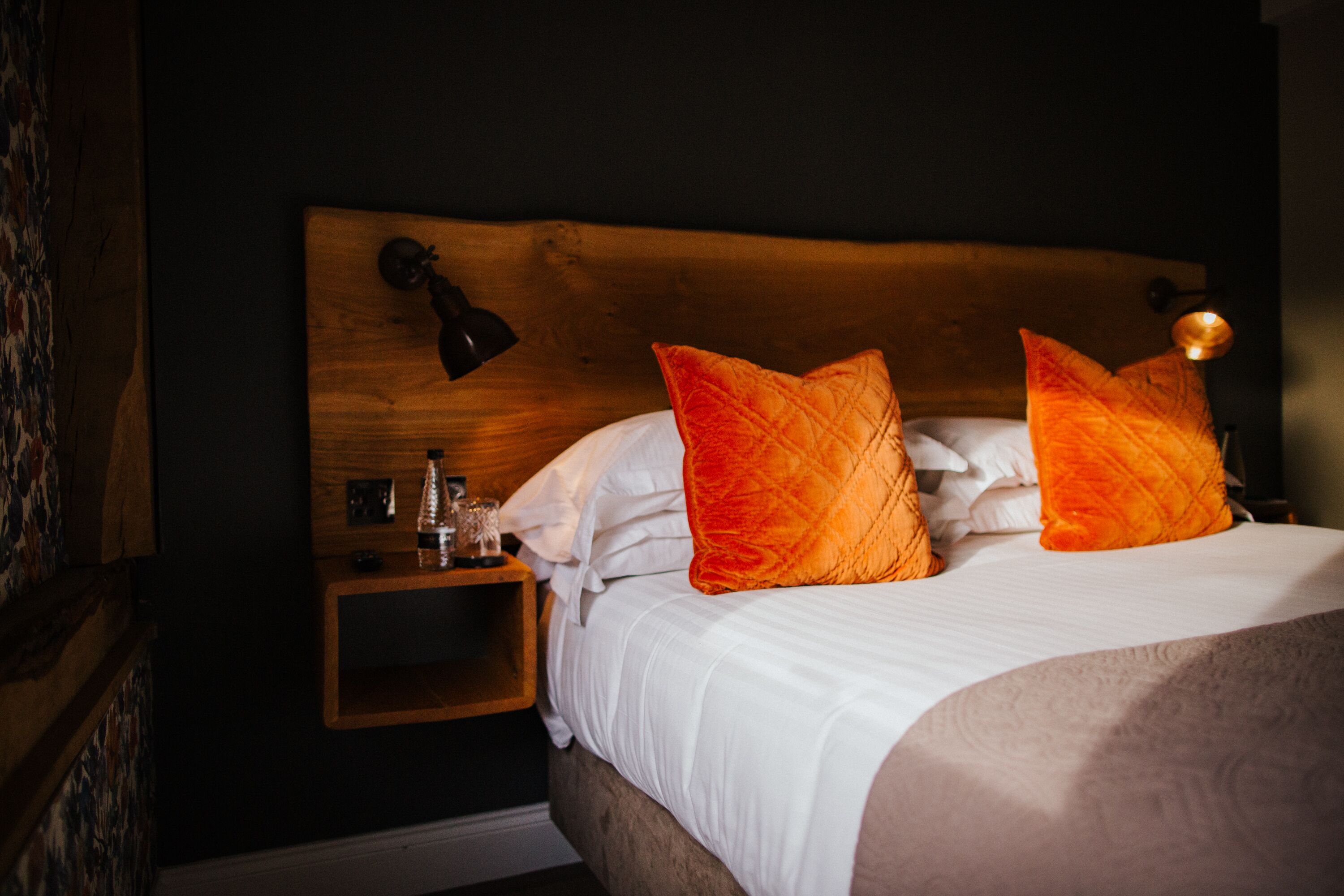 A neatly made bed with white linens and two orange pillows, a wooden headboard, a bedside table with a lamp, and a small shelf with a bottle and glasses.