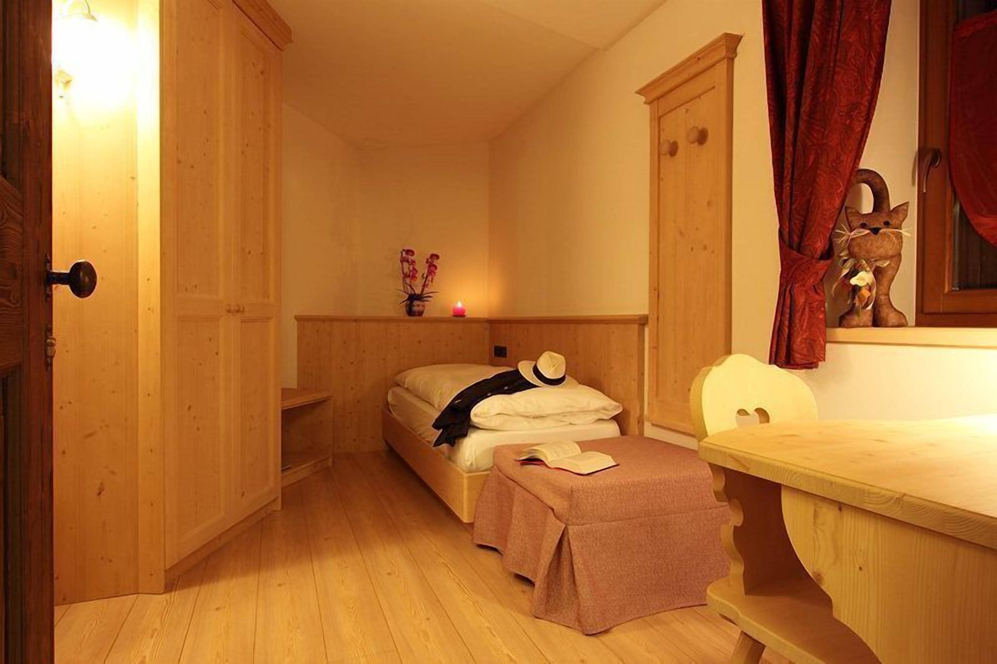 A small, cozy room with wooden walls and a single bed. There is a small table and a chair, and a door is visible on the left.