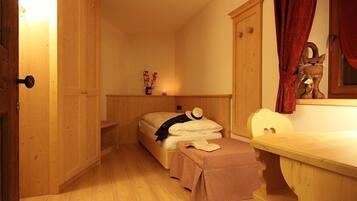 A small, cozy room with wooden walls and a single bed. There is a small table and a chair, and a door is visible on the left.