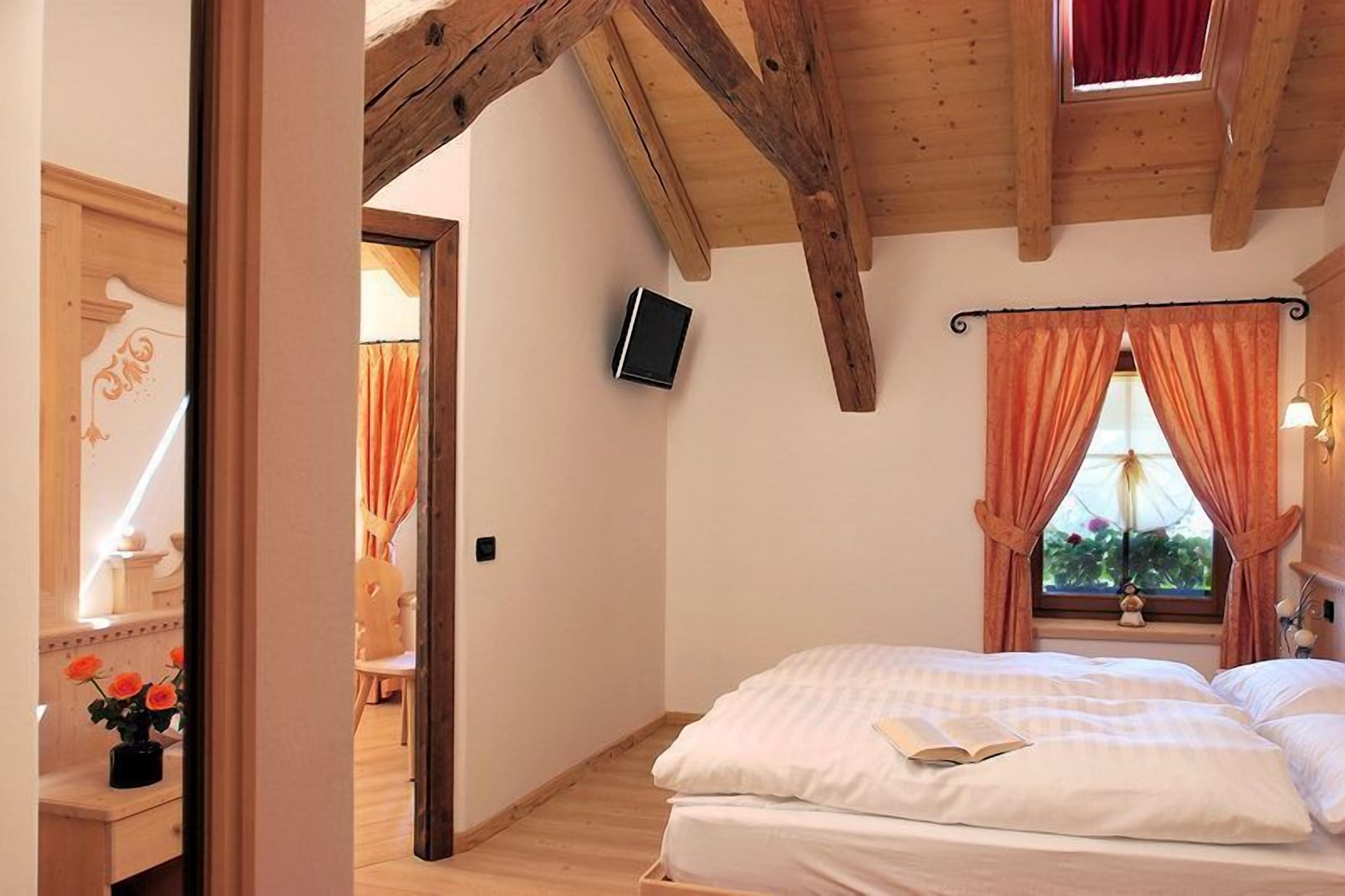 Un dormitorio con techo de madera, cama con ropa blanca, televisor montado en la pared y ventana con cortinas.