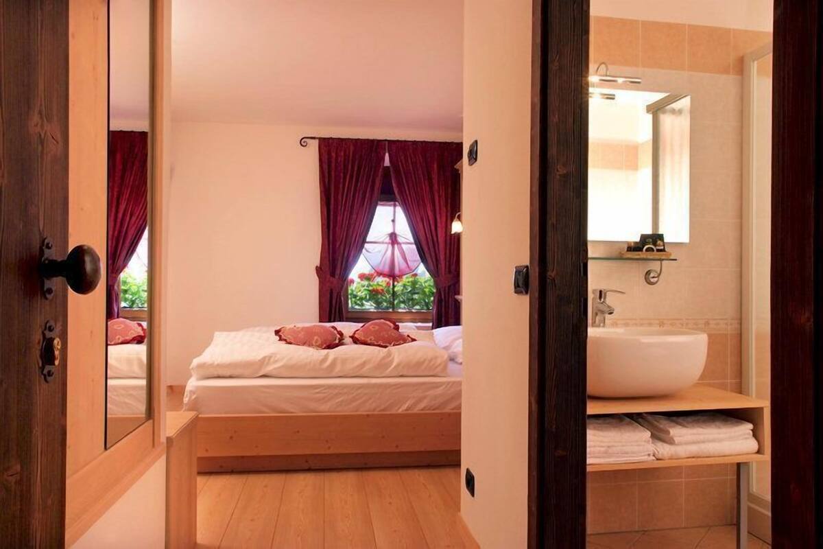 A bedroom with a bed, a window with curtains, a wooden floor, and a bathroom with a sink and mirror.