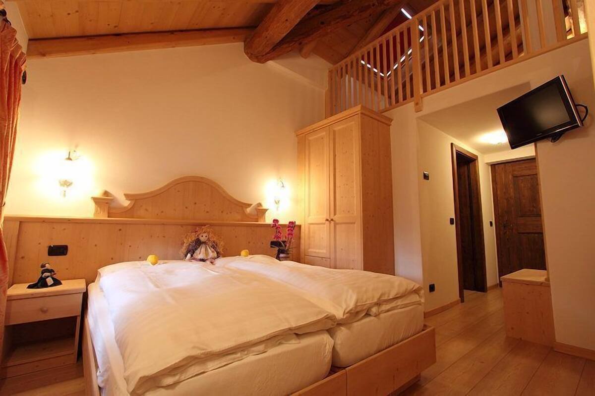 A bedroom with a wooden headboard, a bed with white bedding, a nightstand with a lamp, a wardrobe, and a television mounted on the wall.