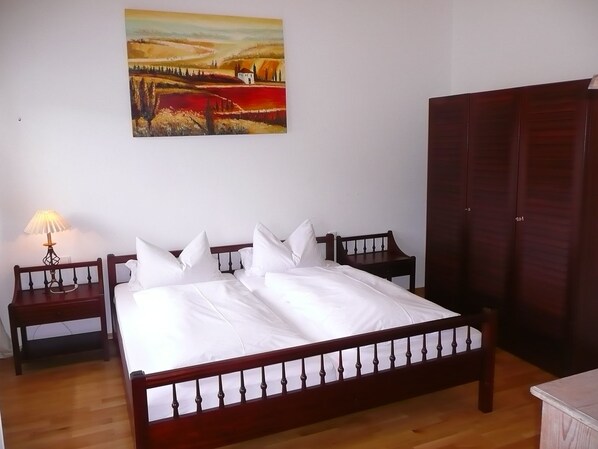 Un dormitorio con cama de madera, ropa de cama blanca, mesita de noche con lámpara, armario y un cuadro en la pared.