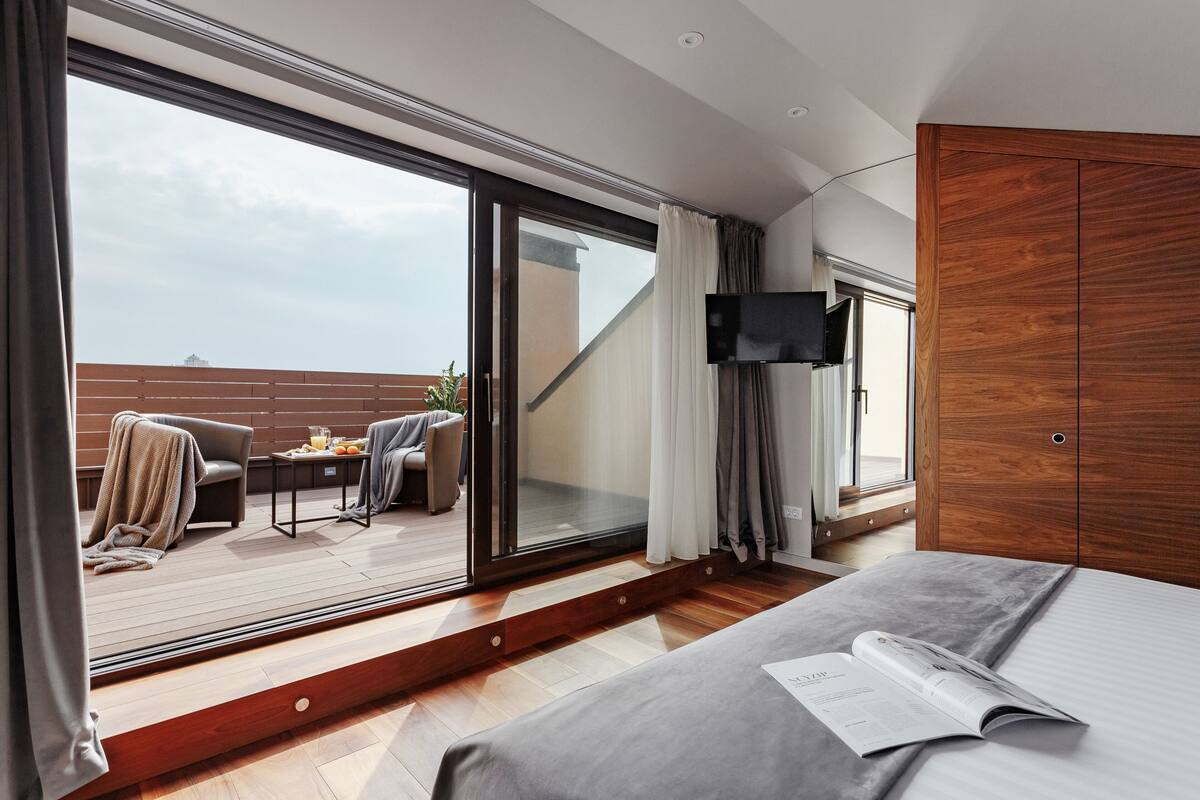 A modern hotel room with a large sliding glass door leading to a balcony, a bed with a magazine, and a TV mounted on the wall.