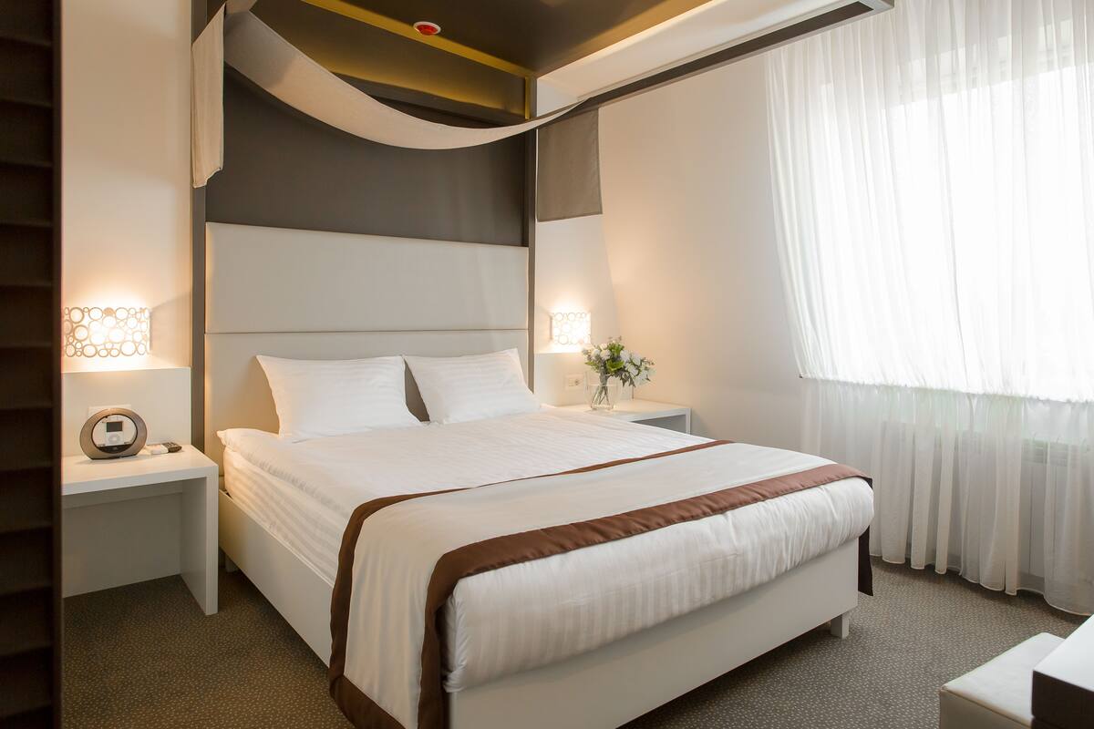 A neatly arranged hotel room with a large bed, bedside tables, and a window with sheer curtains.