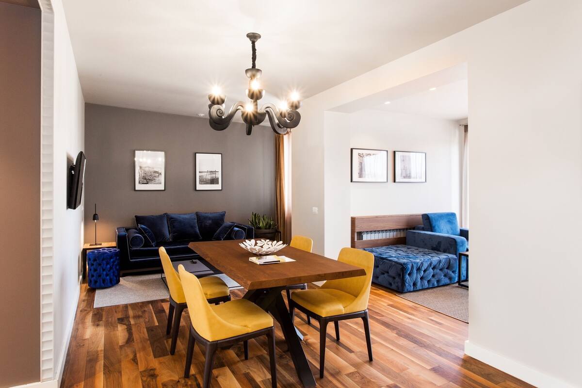 A modern living room with a dining area, featuring a wooden dining table with yellow chairs, a dark sofa, and a blue sofa.