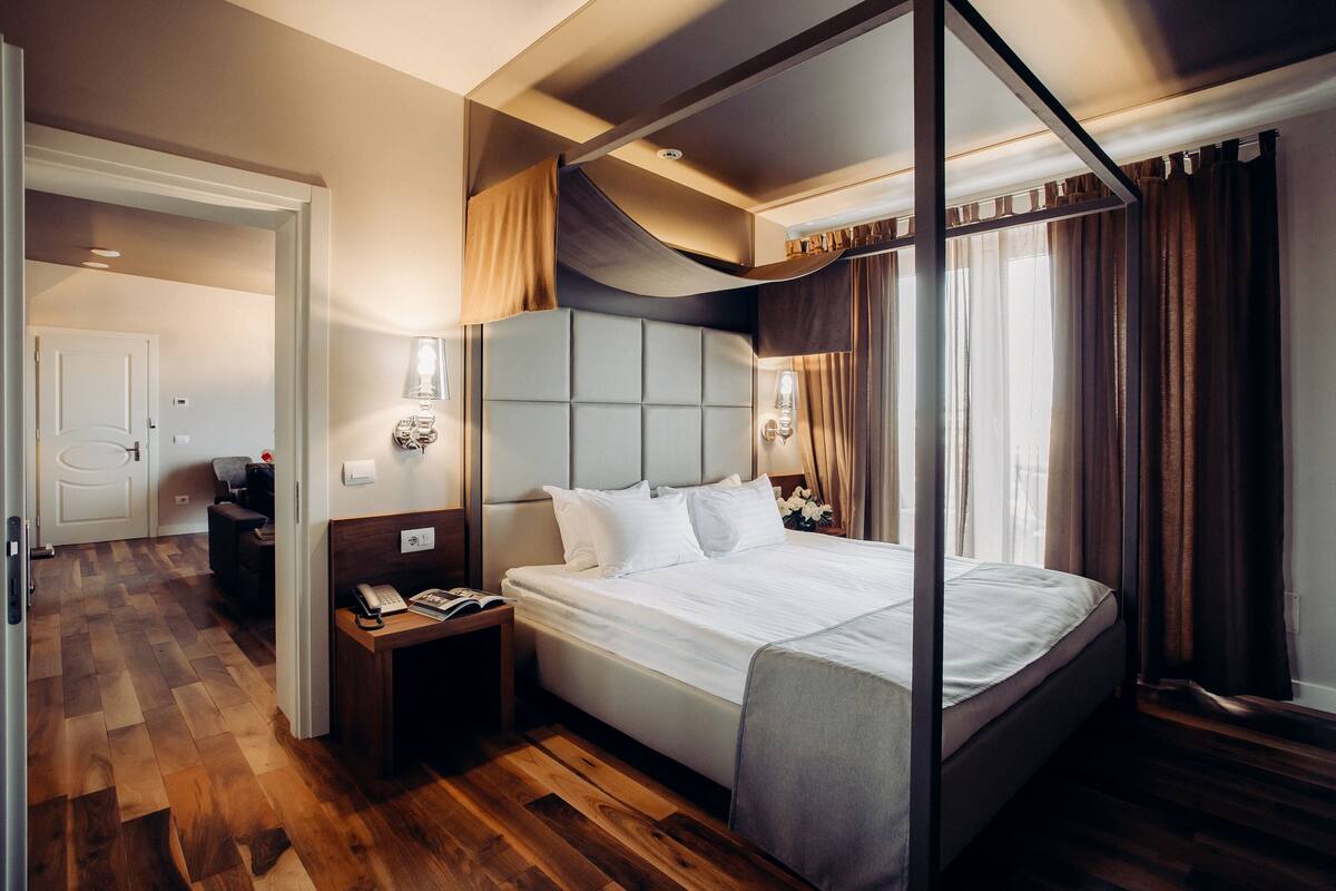 A modern hotel room with a large bed, wooden flooring, a nightstand with a lamp, and a view of the outdoors through a window with curtains.