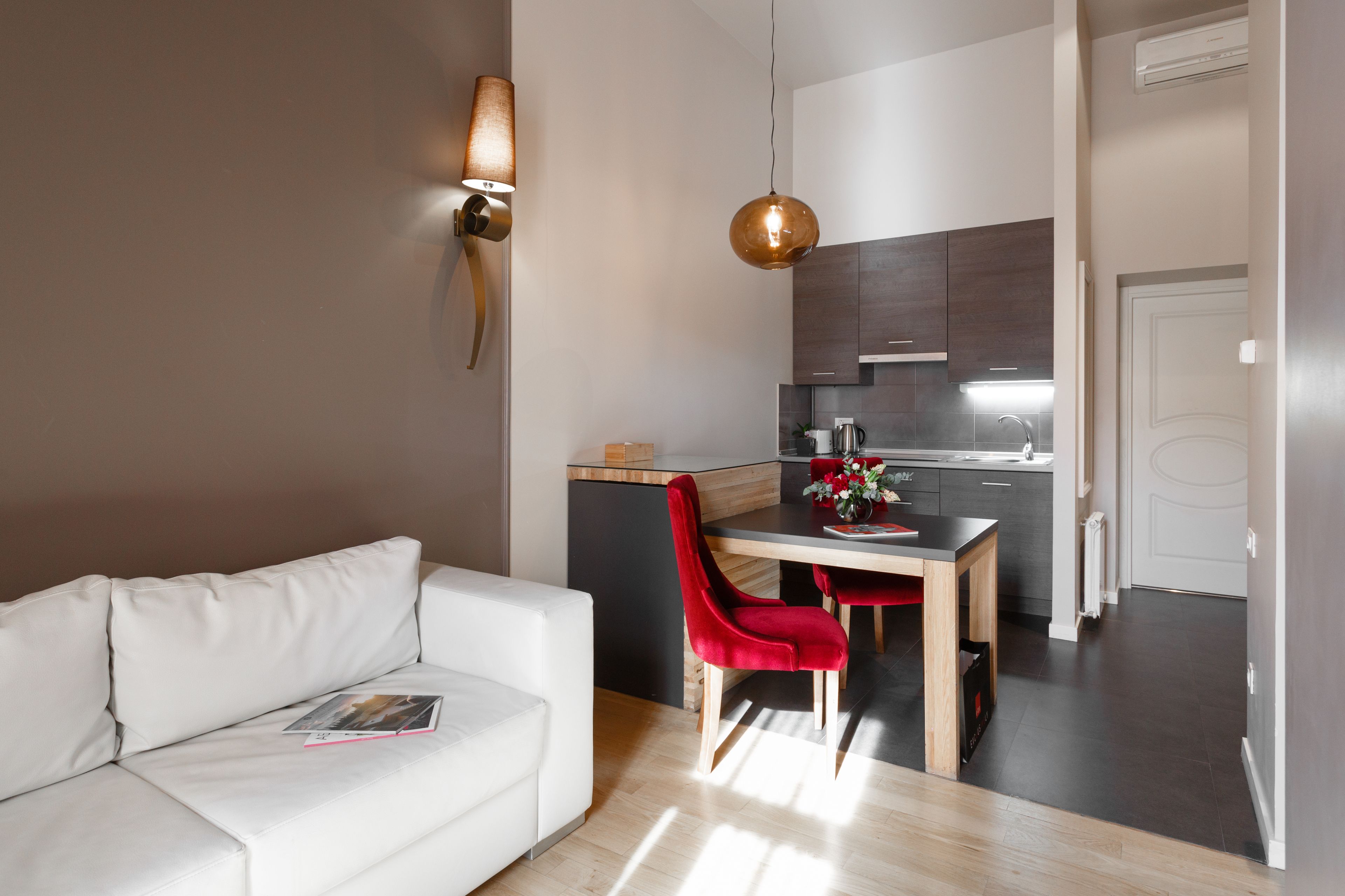 A modern living room with a white sofa, a dining table with red chairs, and a kitchen area with dark cabinets and a hanging light fixture.