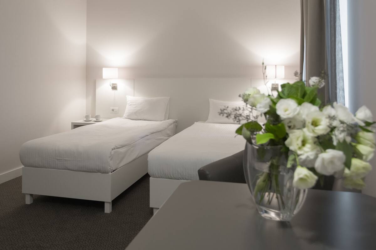 A hotel room with two single beds, a bedside table, a nightstand, a vase with flowers, and a window with curtains.