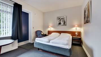 A hotel room with a bed, two bedside tables, a chair, a small table, and a window with curtains.