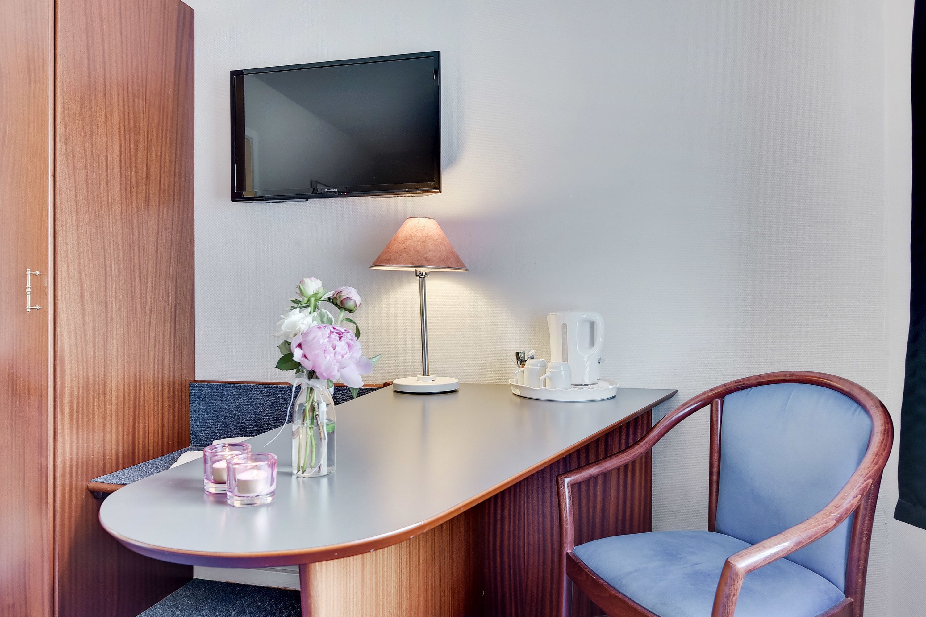 Habitació d'hotel amb escriptori de fusta, cadira blava, televisor fixat a la paret, làmpada, gerra amb flors i cafetera.