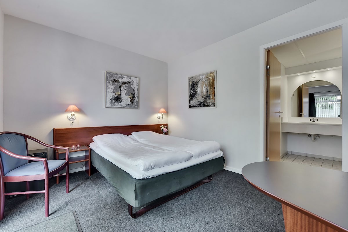 A hotel room with a bed, a chair, a desk, two wall-mounted lamps, and two abstract paintings.