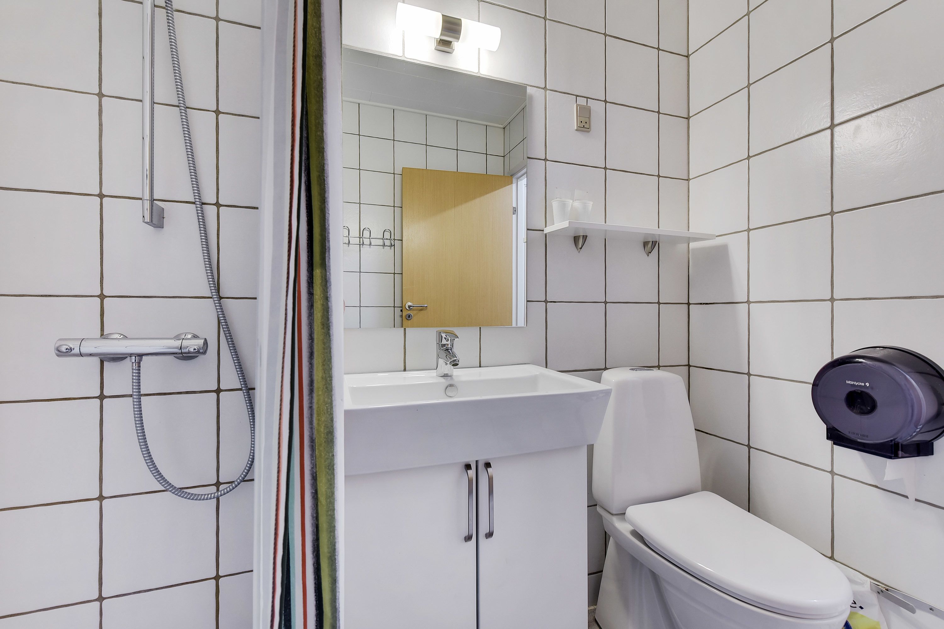 A modern bathroom with a white sink, a toilet, a shower with a handheld showerhead, and a paper towel dispenser.