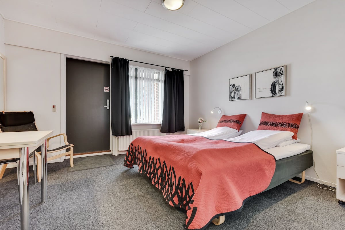 A hotel room with a bed, a desk, and a chair. There is a window with curtains, a wall with framed pictures, and a door.