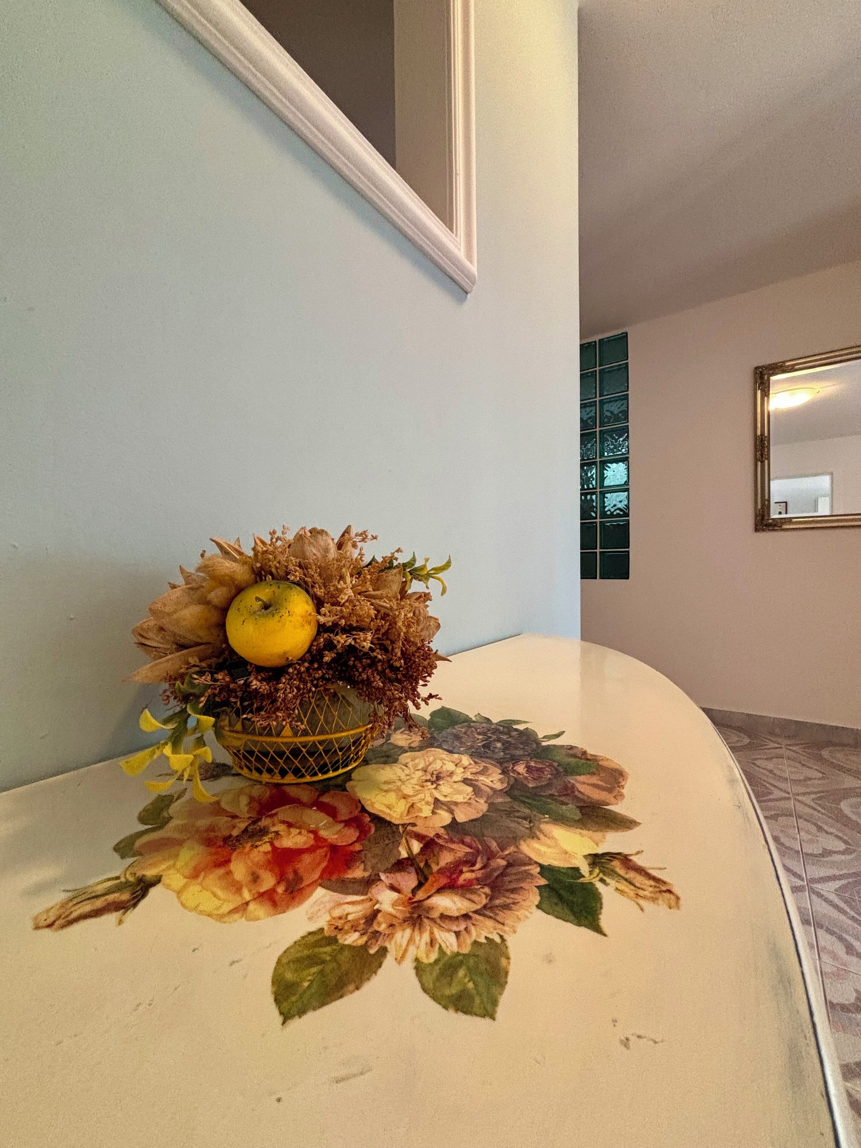 Une table recouverte d’une nappe à motifs floraux, un vase contenant des fleurs séchées et un citron, et un miroir accroché au mur.