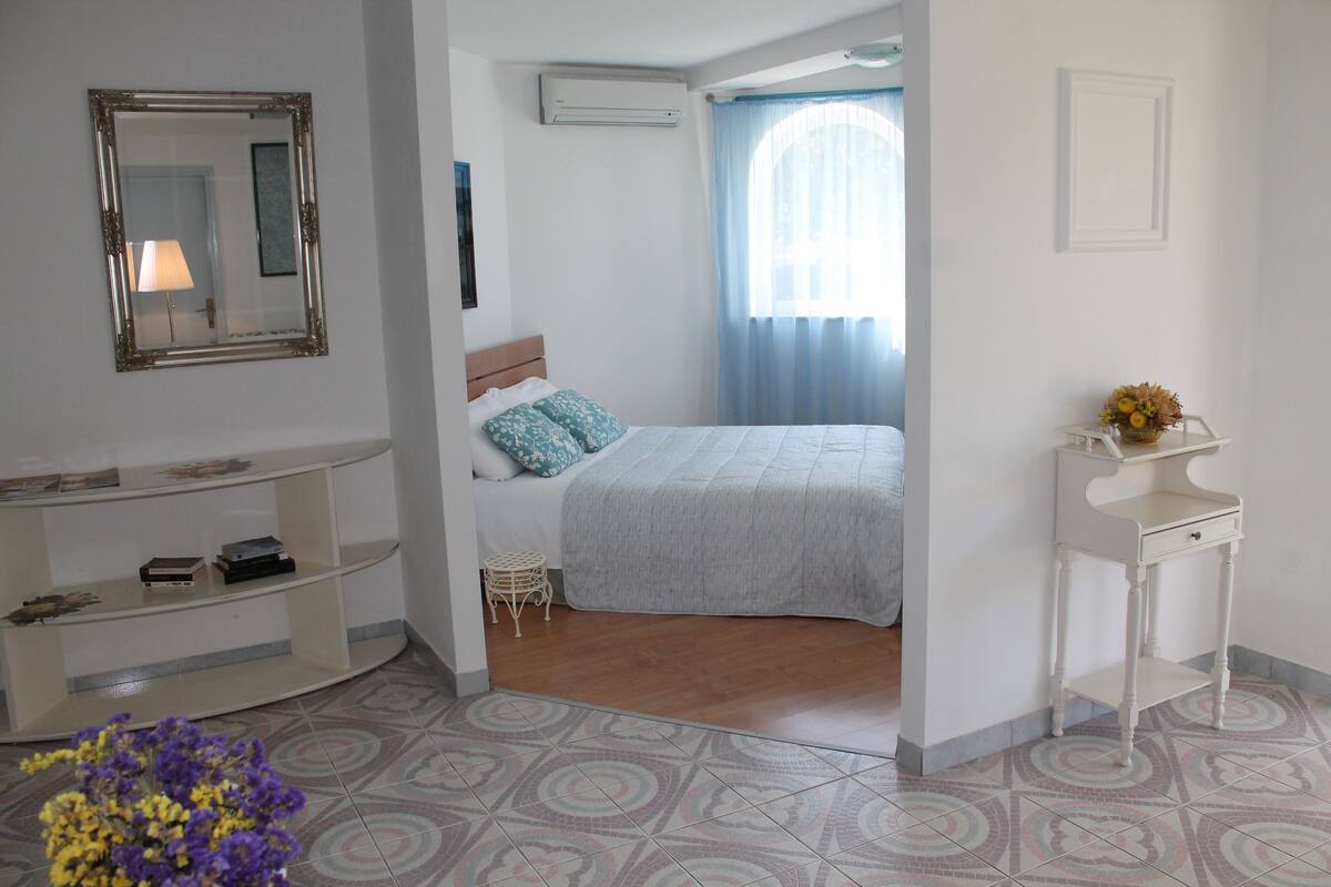 A bedroom with a bed, a bedside table, a mirror, and a small table with a vase.