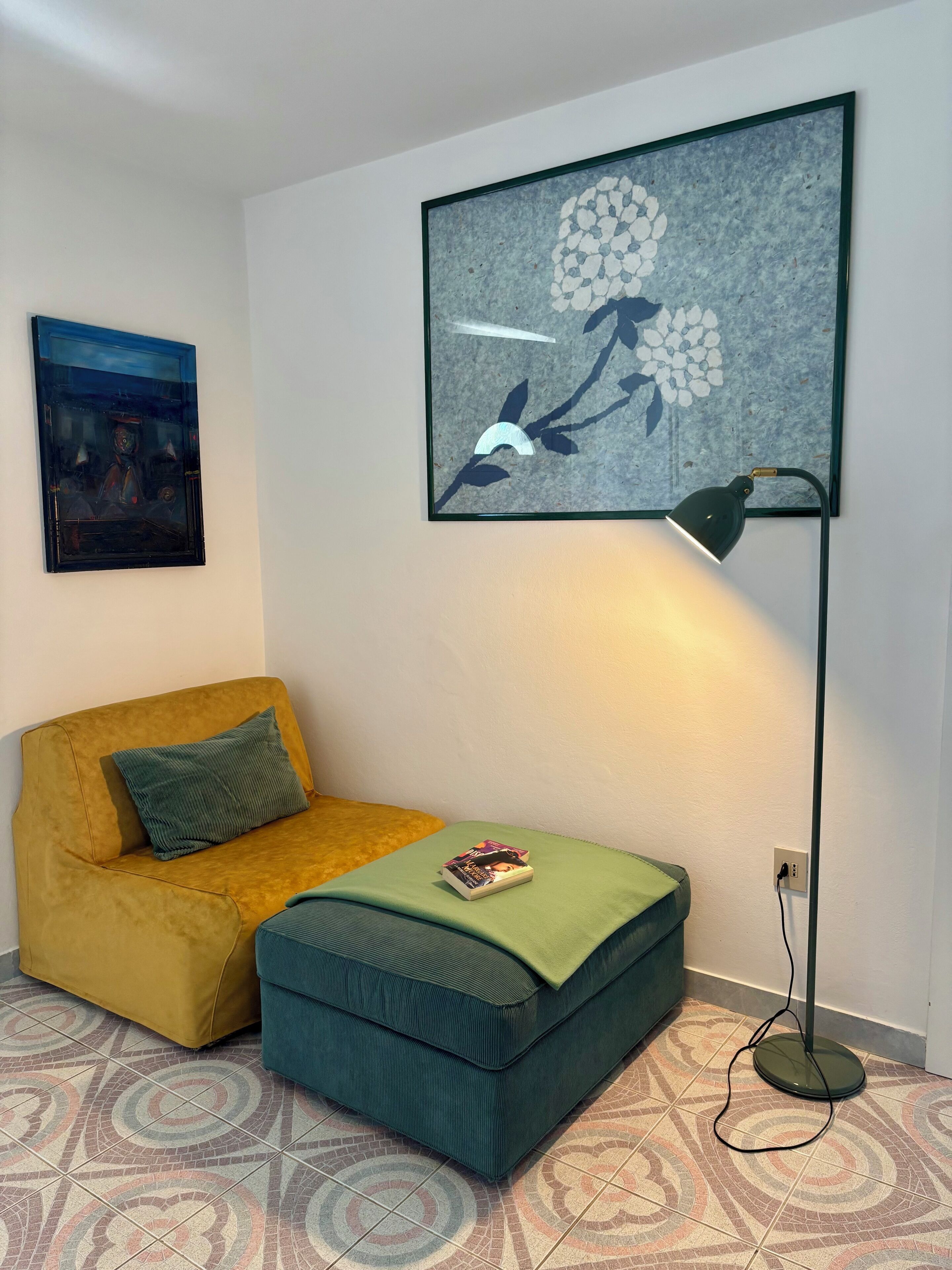 A room with a yellow sofa, a green ottoman, a floor lamp, and a framed abstract painting on the wall.