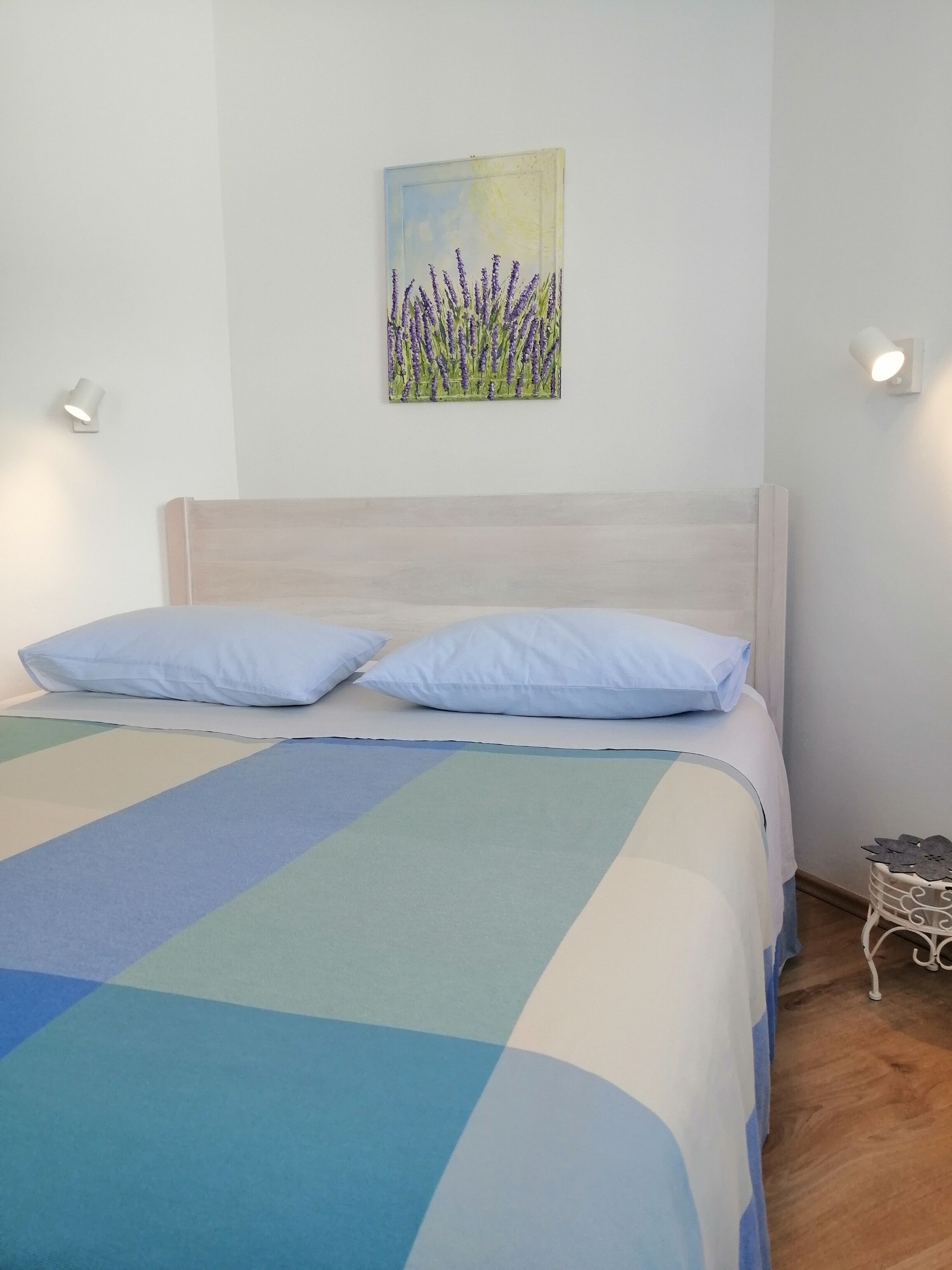 A bed with a blue and green checkered bedspread, two light blue pillows, and a headboard. There is a wall-mounted painting of lavender flowers above the bed.