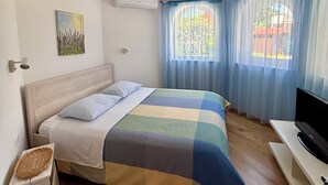 A bedroom with a bed, a TV, a window with curtains, an air conditioning unit, and a wall-mounted artwork. - Villa Mandolina Apartments (Zadar)