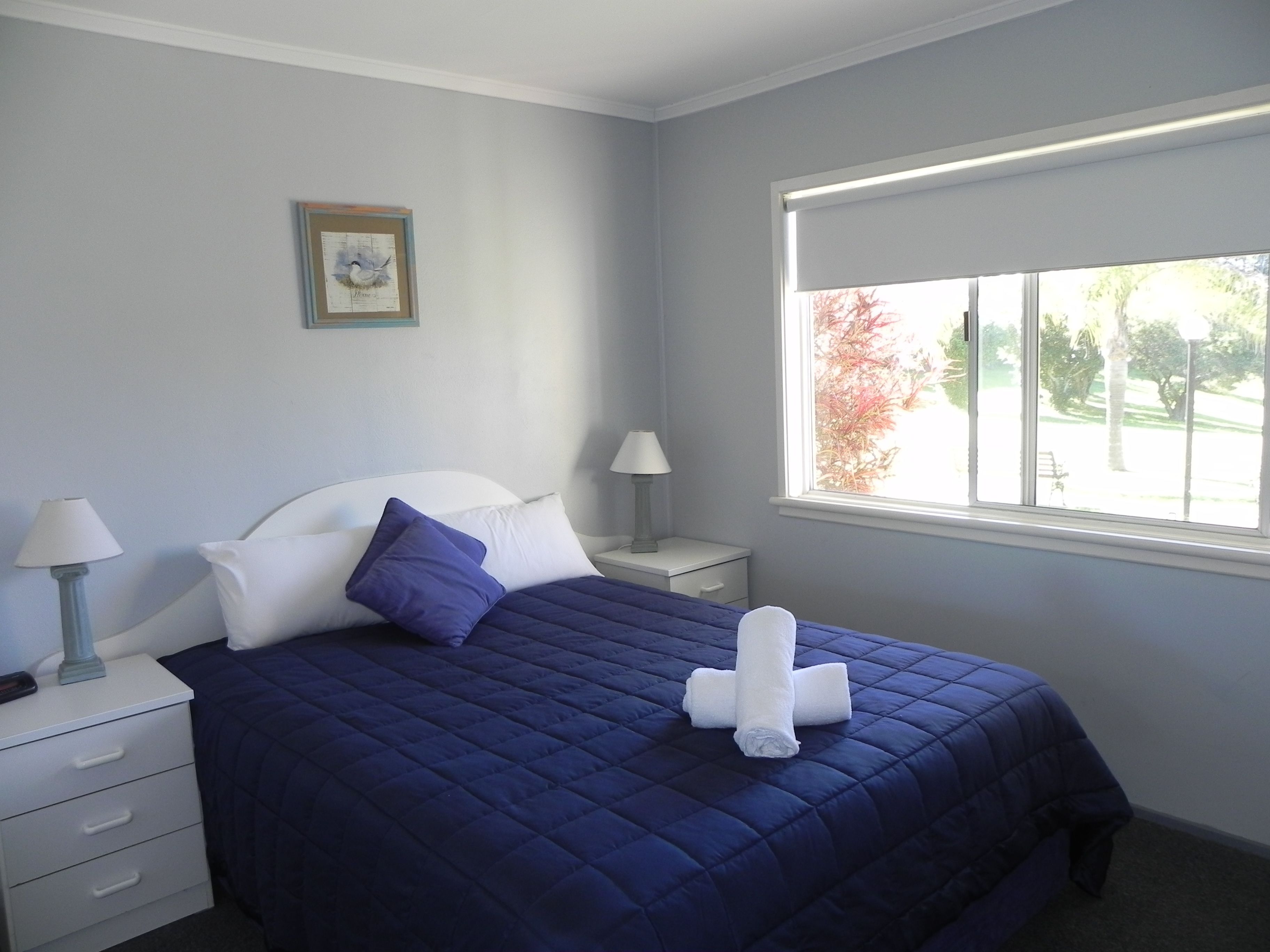 Una cama bien hecha con un edredón azul, almohadas blancas y toallas enrolladas sobre una mesita de noche blanca.