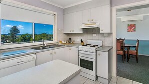 A compact kitchen with white cabinets, a stove, and a sink. There's a window with a view of greenery and a dining area with a table and chairs.