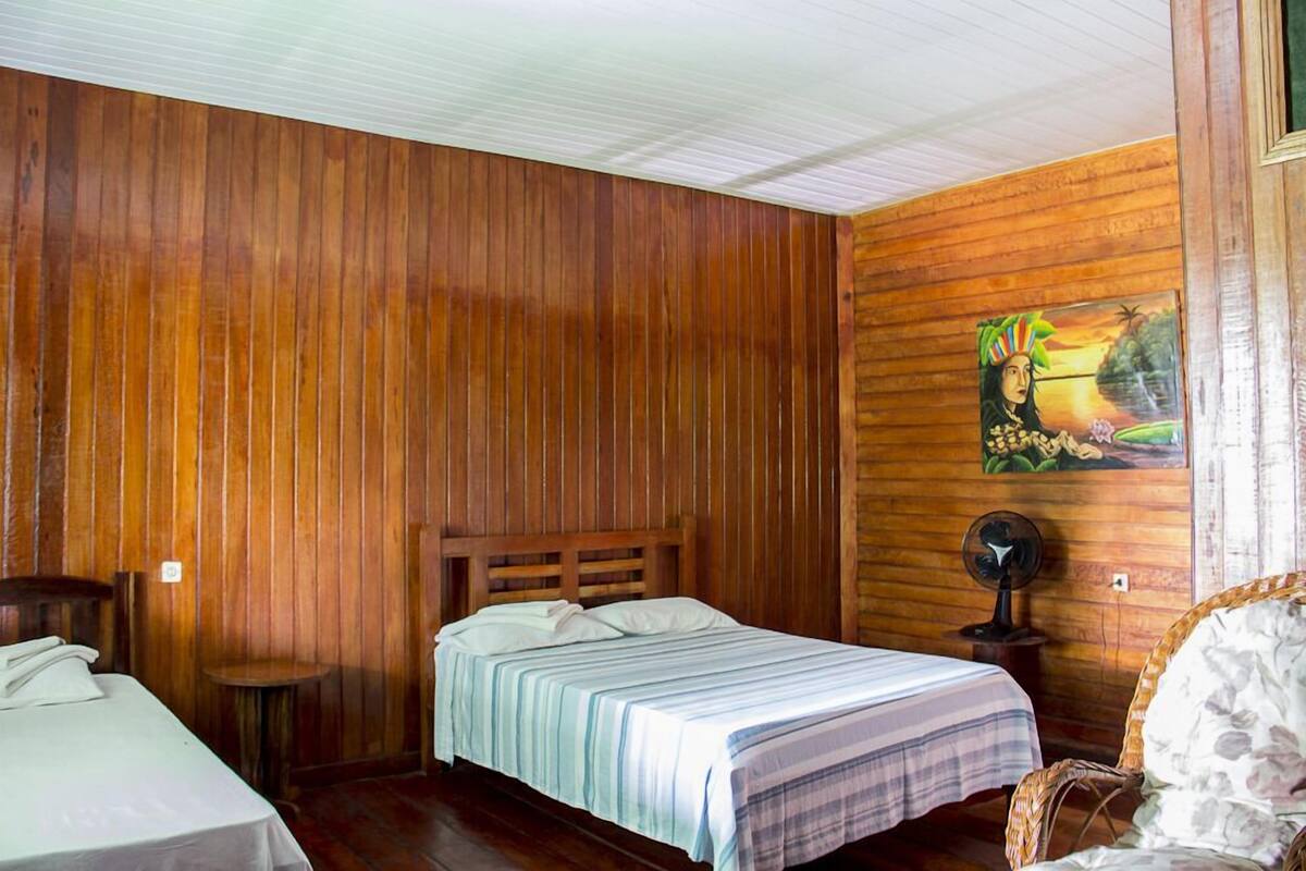 Una habitación con paneles de madera, una cama con colcha a rayas, una pequeña mesa redonda, una silla y un ventilador fijado a la pared.