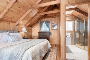 Un dormitorio de cabaña de madera con una cama, mesita de noche, cómoda y un baño visible a través de una puerta de cristal.