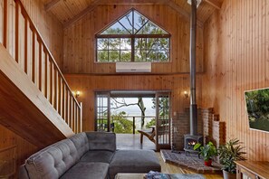A wooden cabin interior with a large window, a fireplace, a sofa, and a dining area. - The Polish Place (Tamborine Mountain)