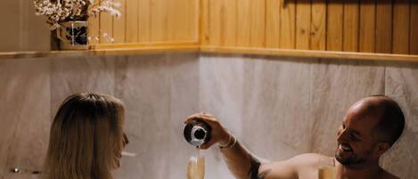 A couple in a bathtub with a camera, surrounded by wooden paneling and a painting.