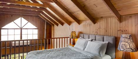 Un dormitorio con una cama grande, vigas de madera en el techo, una mesita de noche con lámpara y una ventana con vistas a la vegetación.