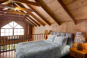 Un dormitorio con una cama grande, vigas de madera en el techo, una mesita de noche con lámpara y una ventana con vistas a la vegetación.
