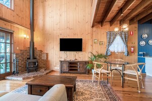A cozy wooden interior with a fireplace, a flat-screen TV, a dining area with chairs, and a large window.