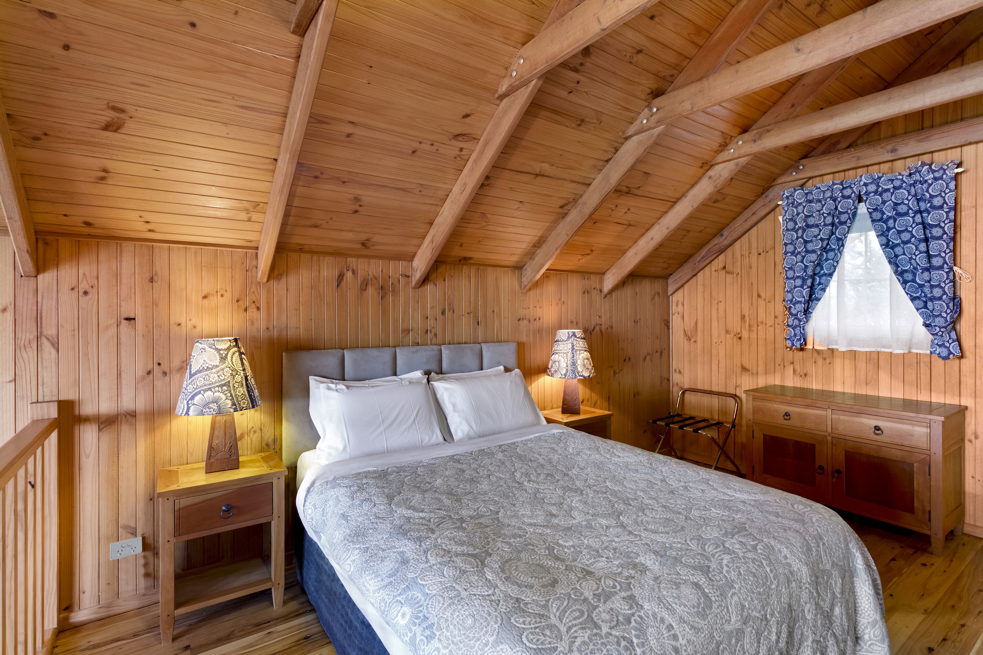 Une chambre à coucher dans une cabane en bois, avec un lit, des tables de chevet, une commode et une fenêtre ornée de rideaux à motifs.