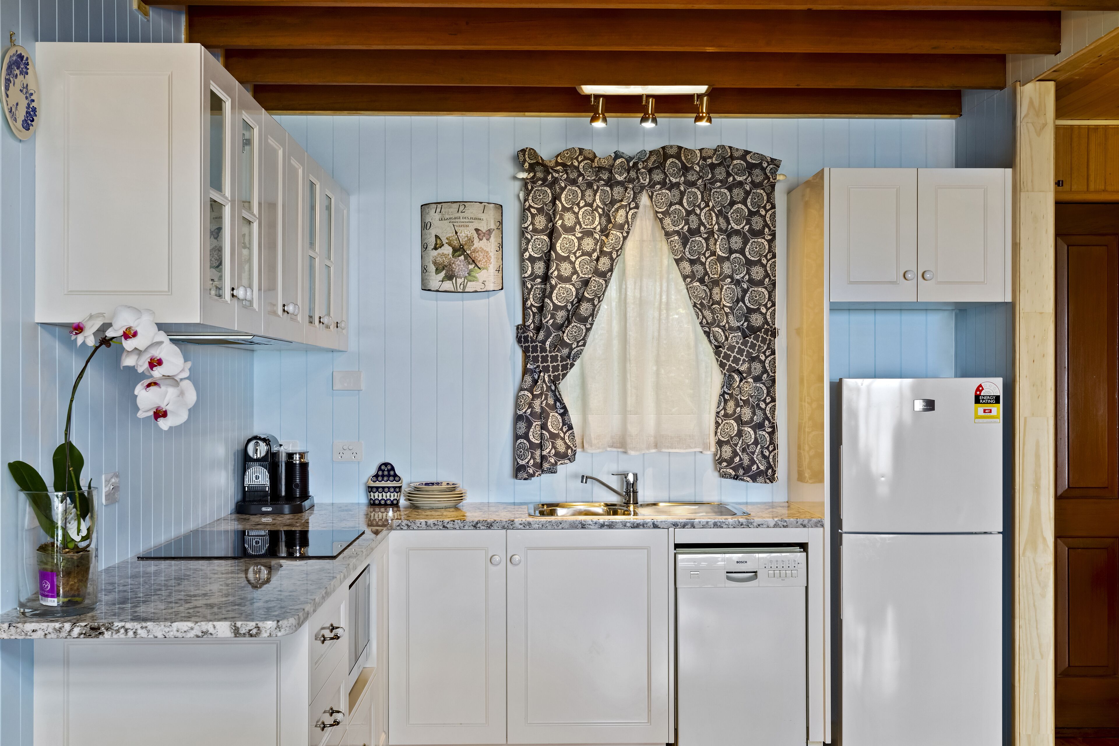 A kitchen with white cabinets, a built-in dishwasher, a refrigerator, a patterned window valance, and a clock on the wall.