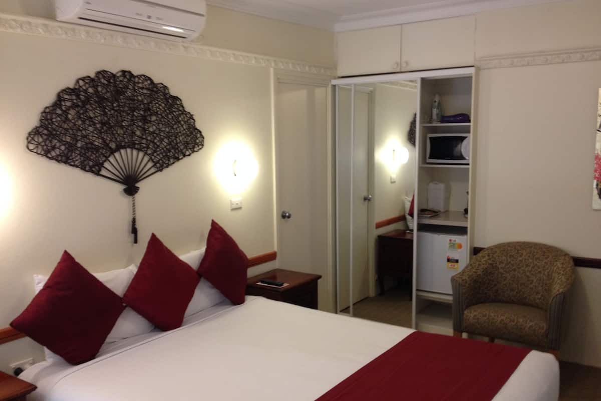 A hotel room with a bed, red pillows, a nightstand, a chair, a wardrobe, and a wall fan.