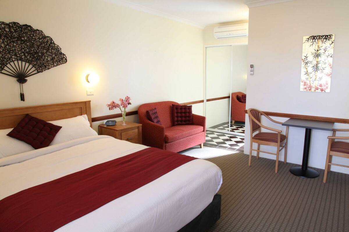 A hotel room with a large bed, a red sofa, a desk with two chairs, a wall fan, and a mirror.