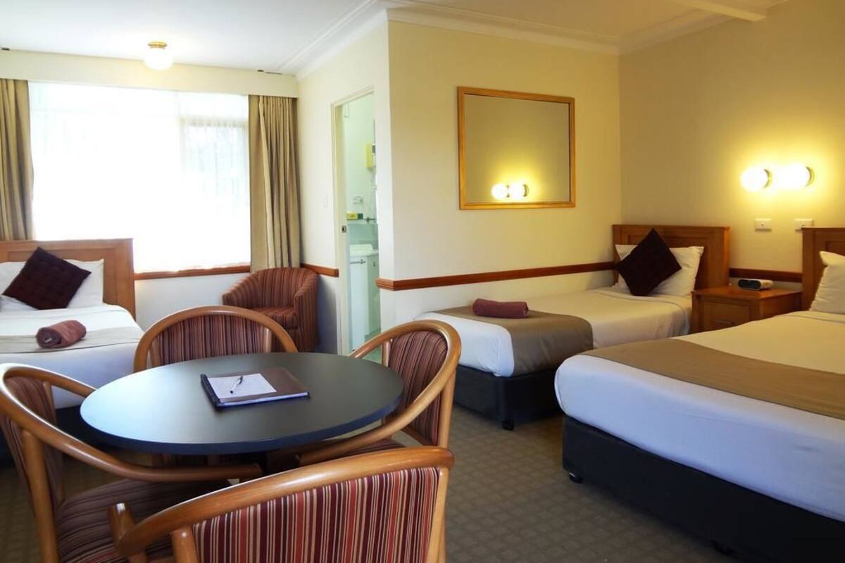 A hotel room with two beds, a round table, and a chair. There is a mirror on the wall and a window with curtains.