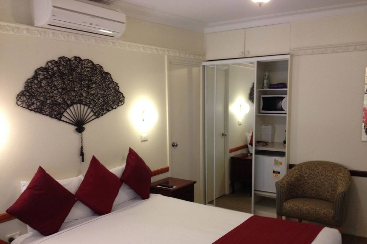 A hotel room with a bed, red pillows, a bedside table, a chair, a wardrobe, and a wall fan.