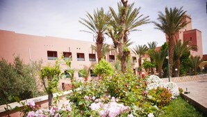 A courtyard with pink flowers, palm trees, and a building with balconies. - Hôtel Saghro (Tinghir)