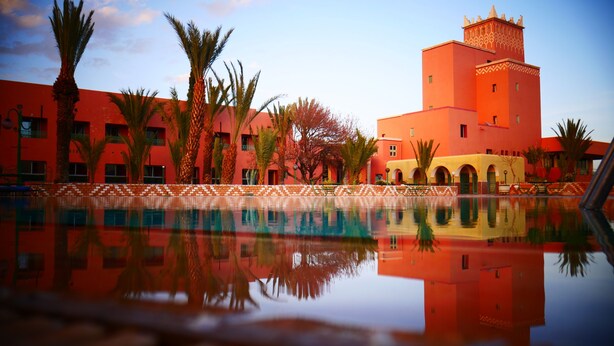 A pool with a reflection of a building with red and yellow walls, palm trees, and a tower. - Hôtel Saghro (Tinghir)
