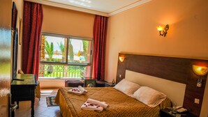 A hotel room with a bed, a desk, a window with curtains, and a view of greenery outside. - Hôtel Saghro (Tinghir)