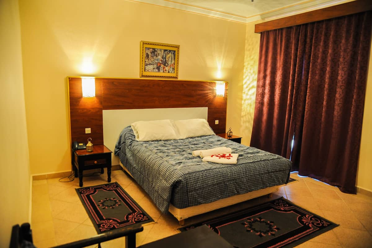A hotel room with a wooden headboard, a bed with a checkered bedspread, two bedside tables with lamps, a nightstand, and a painting on the wall.