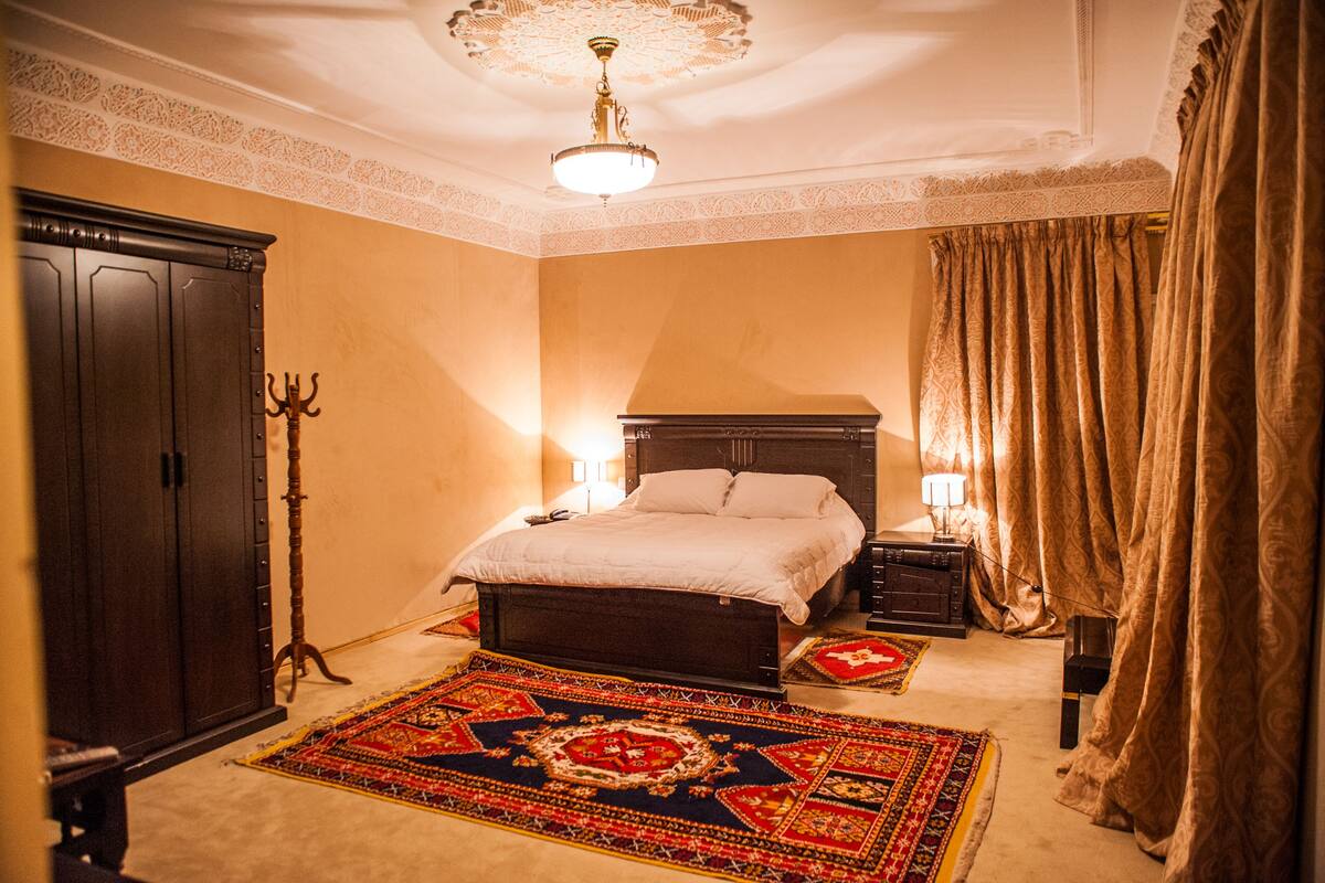 A bedroom with a large bed, a wooden wardrobe, a candelabra, and a patterned rug.