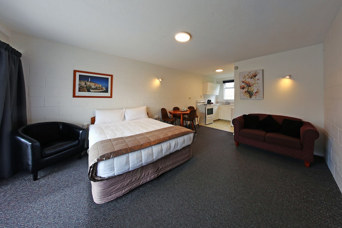 A hotel room with a large bed, a black armchair, a red sofa, a dining table with chairs, and a kitchen area.