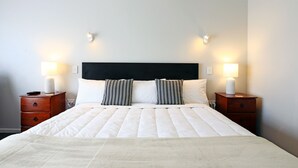 Ein ordentlich bezogenes Bett mit weißer Steppdecke, zwei gestreifte Kissen und einem schwarzen Kopfteil. Auf den beiden Nachttischen stehen Lampen, und auf einem liegt eine Fernbedienung.